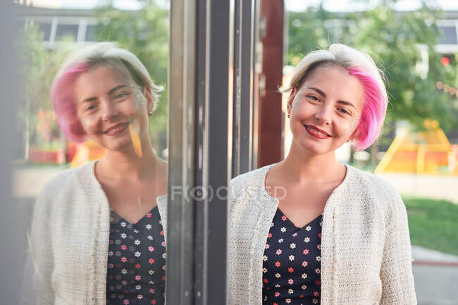 Positive alternative female with dyed hair standing near glass mirrored wall in street and looking at camera — Stock Photo