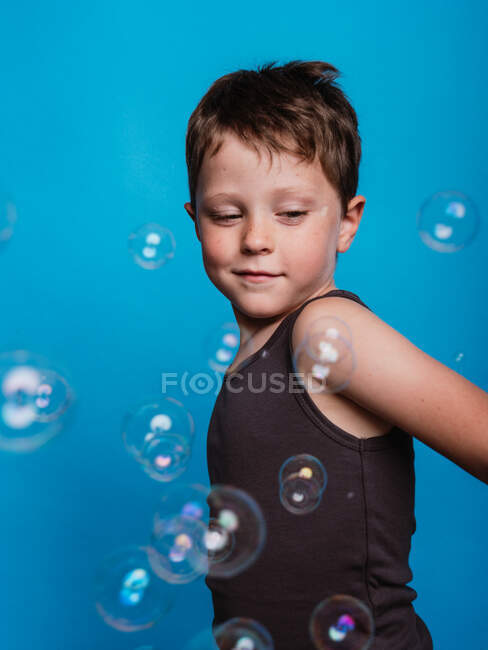 Preteen boy looking away in studio with flying soap bubbles on blue background — Stock Photo