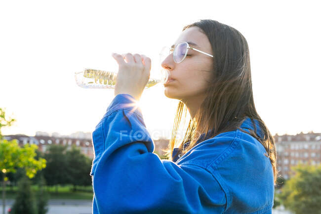 Vista lateral de hembra sedienta con los ojos cerrados bebiendo agua fresca de la botella de plástico en la ciudad en la parte posterior iluminada - foto de stock