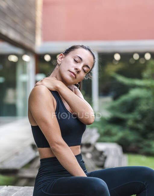 Молодая спортсменка с закрытыми глазами трогает и растягивает шею и плечо во время тренировки во дворе — стоковое фото