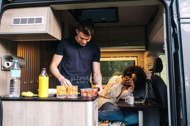 Young African American woman with smartphone taking photo of boyfriend preparing healthy meal inside camper van during summer holidays together — Stock Photo