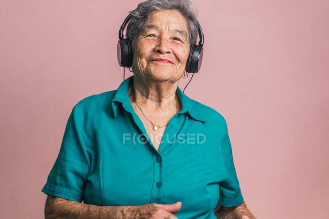 Радісна сучасна старша жінка слухає музику в навушниках і танцює із закритими очима на рожевому фоні в студії — стокове фото