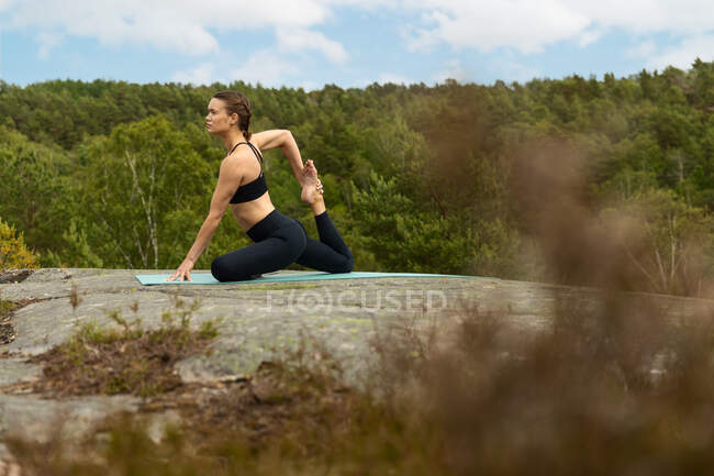 Slim femmina a piedi nudi in nero activewear stretching corpo in Eka Pada Raja Kapotasana posa sulla pietra durante la sessione di yoga in campagna — Foto stock