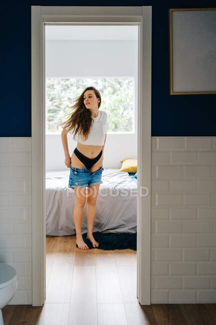 Charmante schlanke Frau zieht Jeanshosen an, während sie zu Hause im Schlafzimmer steht — Stockfoto