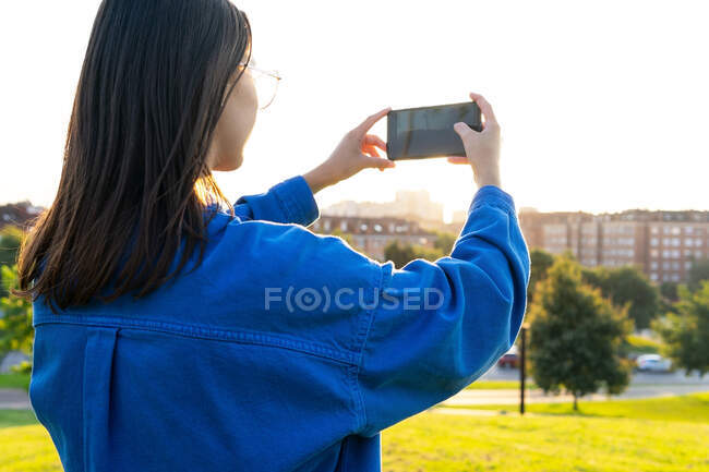 Vista posterior de la mujer con estilo de pie en la colina cubierta de hierba verde y tomar una foto del paisaje urbano en el teléfono inteligente en el día soleado - foto de stock