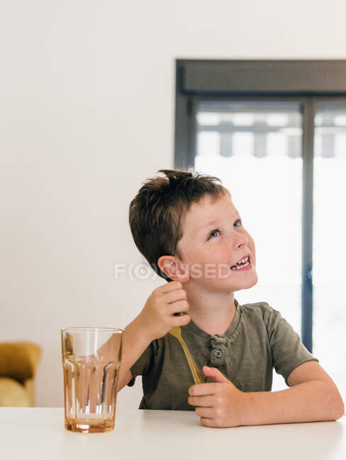Cute preteen child playing with spoon while sitting at table in kitchen and looking away — Stock Photo