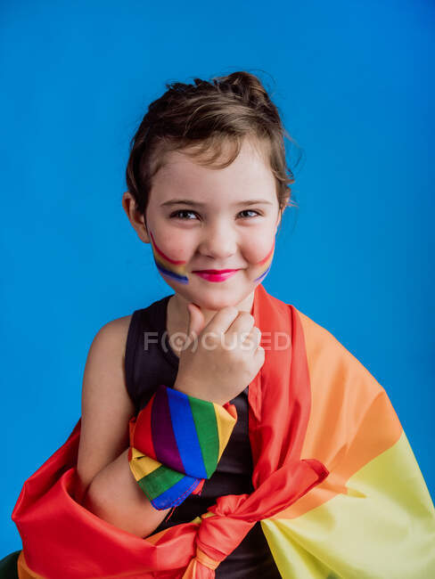 Chica sonriente con mejilla pintada con bandera multicolor sobre fondo azul vivo - foto de stock