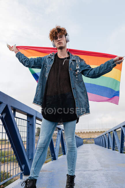 Гомосексуальный стильный мужчина с разноцветным ЛГБТ-флагом, стоящий на мосту и слушающий музыку в наушниках, глядя в камеру — стоковое фото