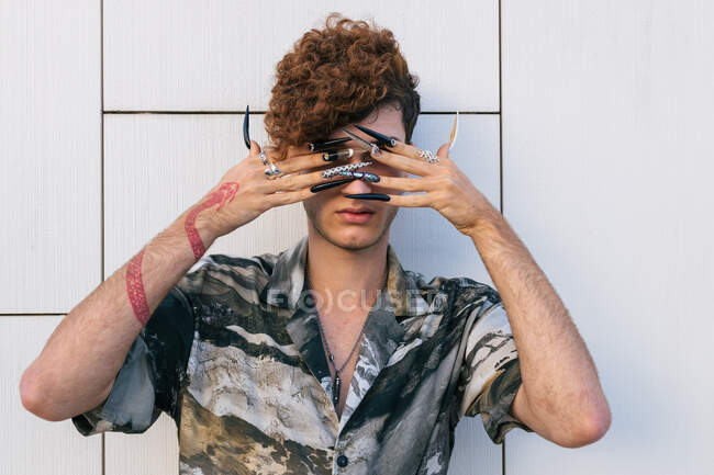 Homem vaidoso jovem em desgaste elegante com unhas compridas em pé na parede de azulejos cobrindo os olhos — Fotografia de Stock