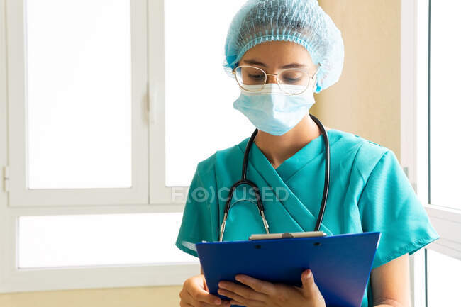 Female medic in protective mask and uniform reading paper medical report on clipboard while standing in hospital — Stock Photo
