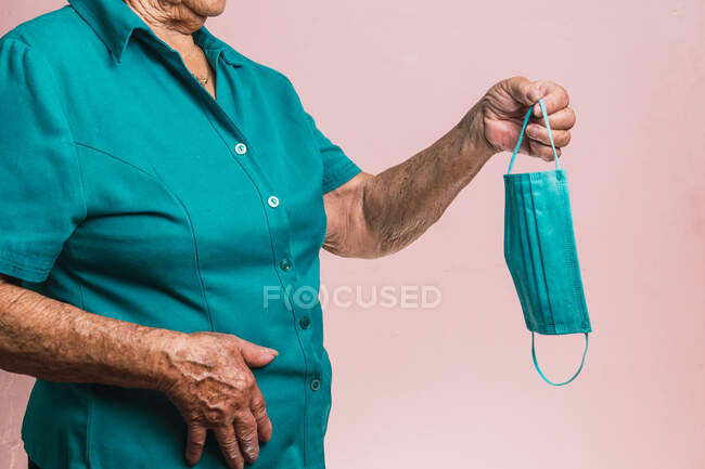 Recadrée femme âgée méconnaissable debout avec un masque médical bleu jetable de COVID et regardant la caméra sur fond rose en studio — Photo de stock
