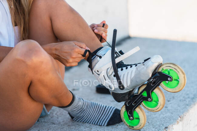 Seitenansicht beschnitten unkenntliche Skaterin sitzt auf dem Boden im Stadtgebiet und setzt auf Rollerblades, während sie das Wochenende im Sommer genießt — Stockfoto