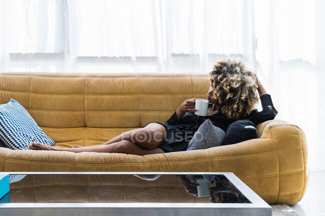 Serene African American female with Afro hair lying with mug of beverage on soft sofa in living room and chilling at weekend at home — Stock Photo