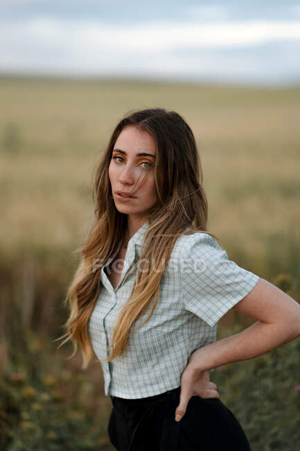 Side view of young mindful female looking at camera on road near meadow under cloudy sky in evening in countryside — Stock Photo