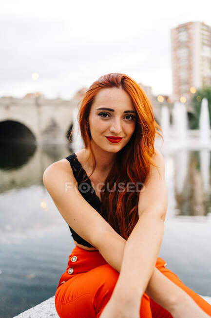 Femme heureuse avec de longs cheveux roux et en pantalon orange vif assis sur la frontière sur la promenade en ville regardant la caméra — Photo de stock