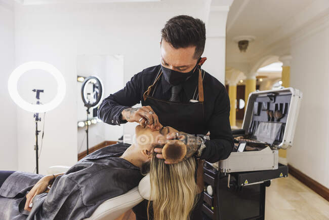 Mann mit Maske beschmiert Gesicht einer blonden Frau bei Arbeit im professionellen Make-up-Studio — Stockfoto