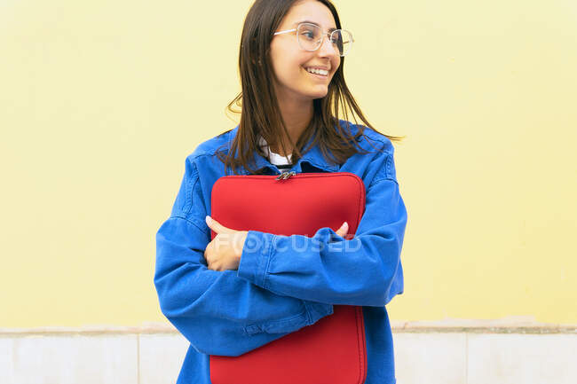 Heureuse pigiste féminine avec netbook dans le cas rouge debout dans la rue de la ville et regardant loin — Photo de stock