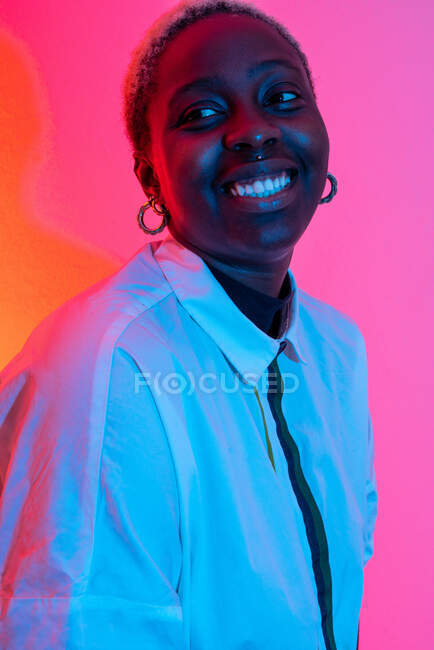 Deliziosa donna afroamericana sorridente in studio con luci al neon colorate e distogliendo lo sguardo — Foto stock