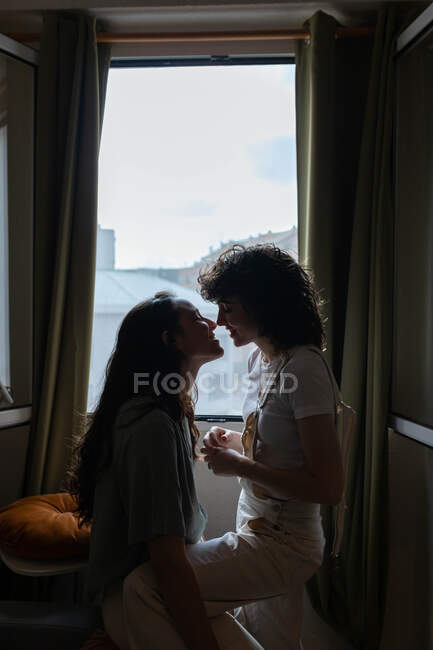 Side view of content LGBT couple of females in love on sofa at home and looking at each other with love — Stock Photo
