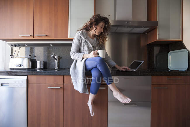 Business woman with curly hair sitting in the kitchen taking an infusion while using her laptop and working at home — Stock Photo