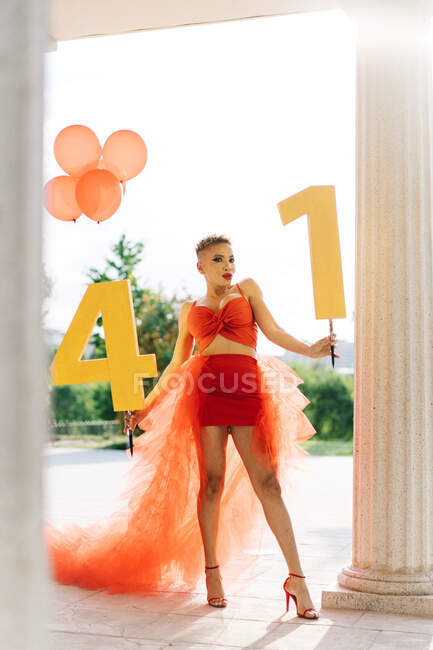 Модная взрослая афроамериканка в красном платье с декоративными номерами и воздушными шарами, смотрящая в камеру на дорожке во время вечеринки — стоковое фото