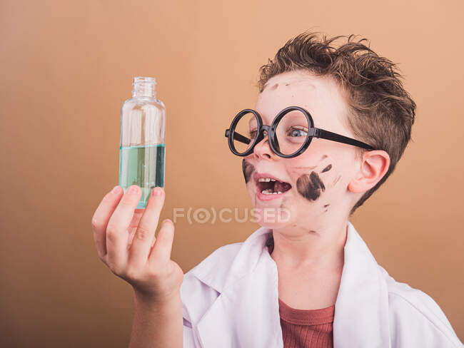 Garoto surpreso em óculos e roupão de laboratório com rosto sujo e líquido em garrafa depois de fazer experiências químicas — Fotografia de Stock