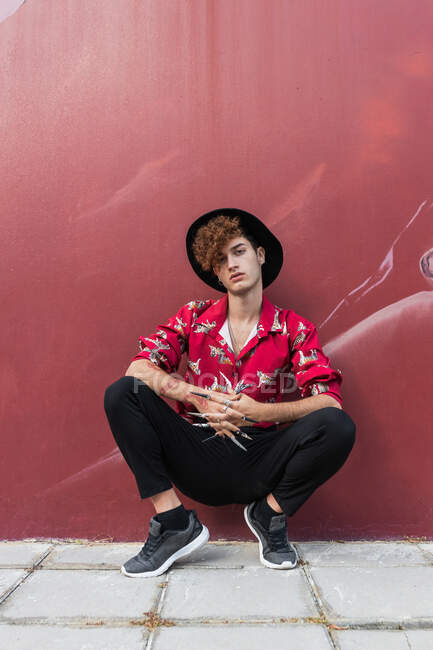 Homem homossexual na moda com unhas compridas em camisa ornamental olhando para a câmera enquanto se agacha no pavimento contra a parede — Fotografia de Stock