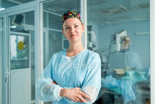 Médico adulto seguro de sí mismo con brazos cruzados en gorra médica ornamental mirando a la cámara contra la pared de vidrio en el hospital - foto de stock