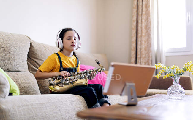 Criança consciente em fones de ouvido com olhos fechados e saxofone no sofá gravação de vídeo no celular em casa — Fotografia de Stock
