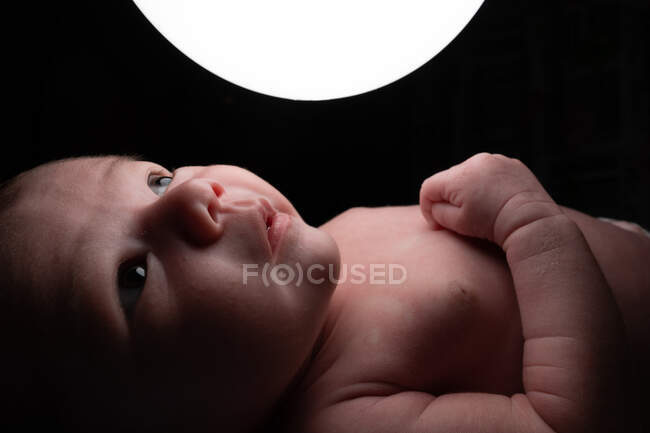 Side view of tender infant on bed and touching glowing night light lamp in dark room — Stock Photo