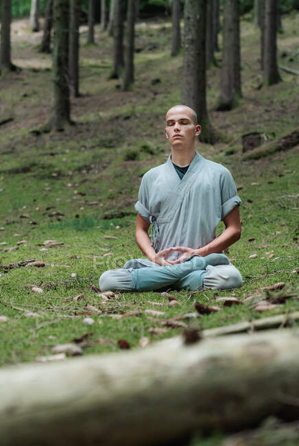 Bald man in traditional clothes sitting on grass in Lotus pose and meditating during kung fu training in forest — Stock Photo