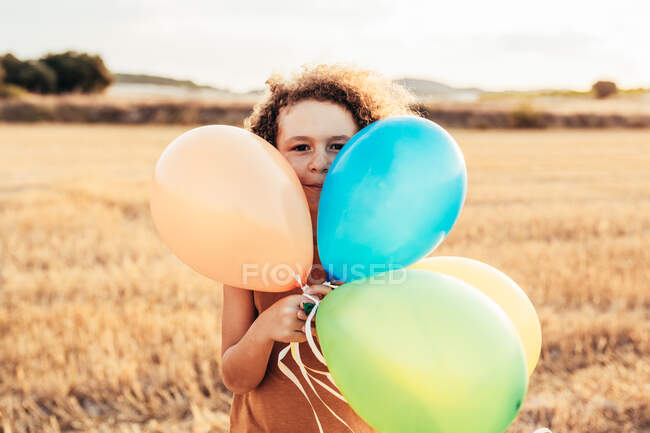 Етнічна дитина з кучерявим волоссям грає з різнокольоровими повітряними кульками в літньому полі і дивиться на камеру — стокове фото