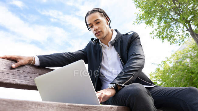 Von unten ein männlicher Freiberufler mit geflochtener Frisur, der auf einer Bank sitzt und auf der Laptop-Tastatur tippt, während er mit TWS-Ohrhörern ferngesteuert an einem Projekt in der Stadt arbeitet — Stockfoto