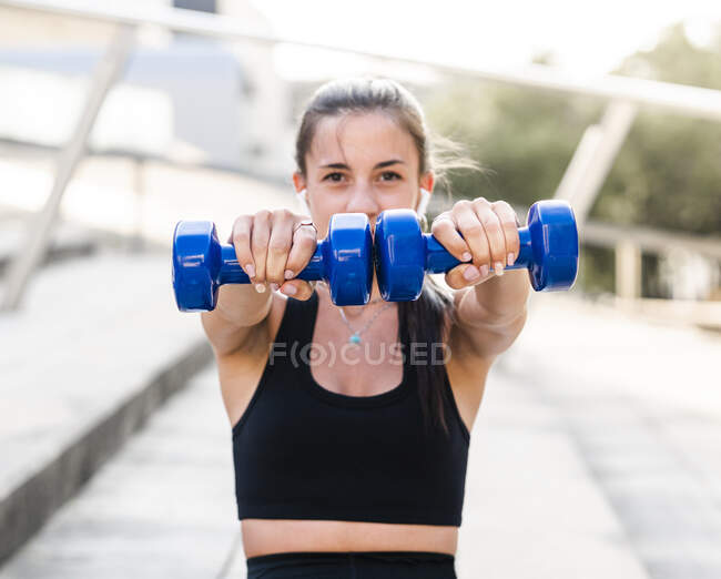 Soft focus of determined female athlete doing exercise with dumbbells during fitness workout on city street in summer — Stock Photo