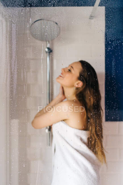 Mujer envuelta en toalla blanca y suave de pie detrás de la puerta de vidrio húmedo de la cabina de ducha y mirando a la cámara - foto de stock