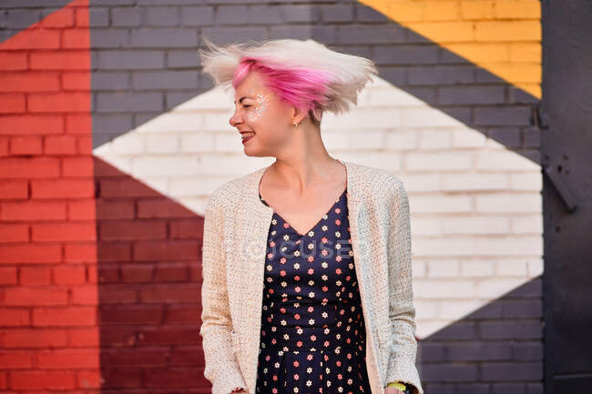 Despreocupado mujer alternativa lanzando cabellos cortos teñidos contra la pared colorida en el área urbana - foto de stock