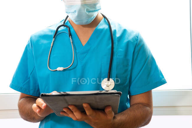 Анонімний чоловічий лікар у формі та стетоскоп стоїть у лікарні та серфінг Інтернету на планшеті — стокове фото