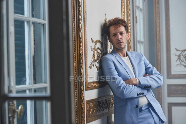 Pensativo hombre de traje de pie con los brazos cruzados y apoyado en la pared en la habitación elegante - foto de stock
