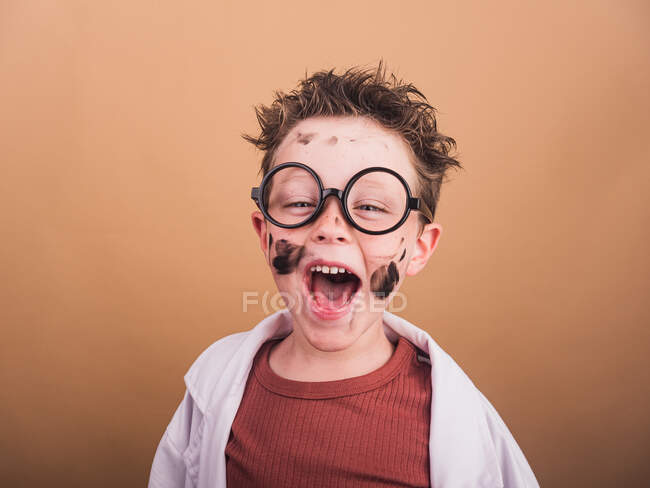 Chemist boy in laboratory robe and plastic glasses looking at camera with mad gaze on beige background — Stock Photo