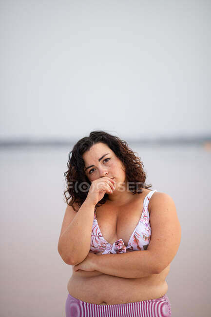 Tranquila mujer curvilínea en bikini de pie sobre fondo de estanque rosa en verano mientras se apoya en la mano y mirando a la cámara - foto de stock