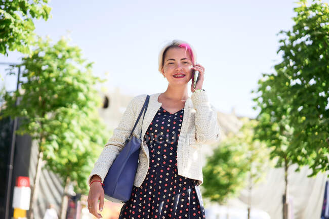 Delighted alternative female with dyed hair standing in street on sunny day in summer and looking at camera while talking on the mobile phone — Stock Photo