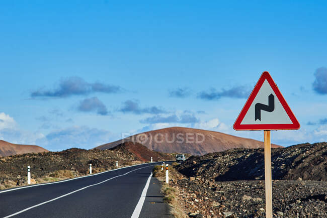 Señal de advertencia de doble curva situada cerca de la autopista de asfalto contra colinas y cielo azul nublado en Fuerteventura, España - foto de stock