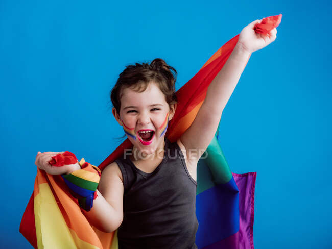 Chica sonriente con mejilla pintada levantando brazos con bandera multicolor sobre fondo azul vivo - foto de stock