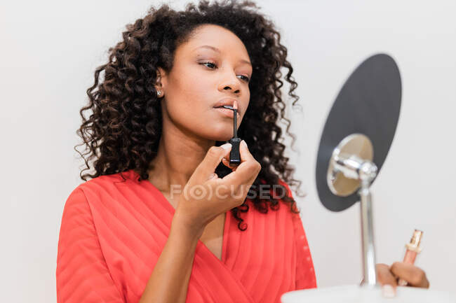 Charming ethnic female in red clothes with curly hair making up lips while looking in mirror on light background — Stock Photo