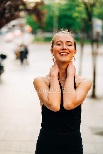 Femme positive dans des vêtements élégants debout n rue de la ville et regardant la caméra tout en touchant le cou — Photo de stock