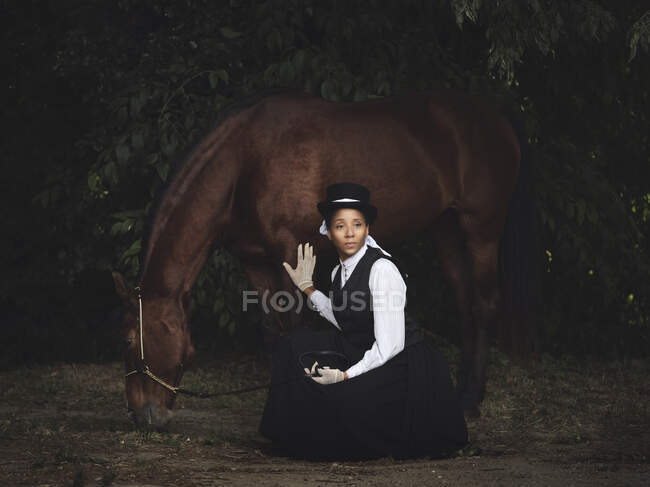 Senhora adulta afro-americana confiante em roupas elegantes e chapéu sentado com cavalo marrom enquanto olha para longe perto de árvores durante o dia — Fotografia de Stock