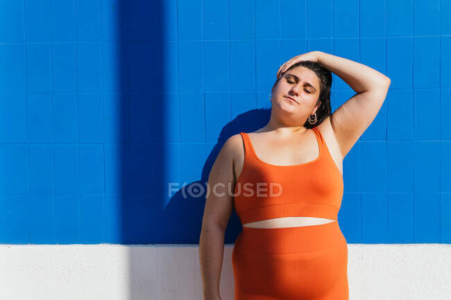 Plus size ethnic female athlete in active wear with closed eyes touching head during training against blue tiled wall in sunlight — Stock Photo
