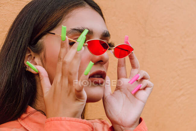 Vista lateral de la hembra carismática del cultivo con uñas largas y brillantes que se ponen gafas de sol de moda contra la pared naranja - foto de stock