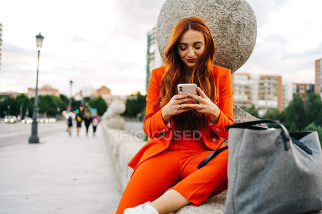 Mujer elegante feliz con el pelo rojo y en traje naranja vibrante sentado en la frontera de piedra en la ciudad y mensajería en el teléfono celular - foto de stock