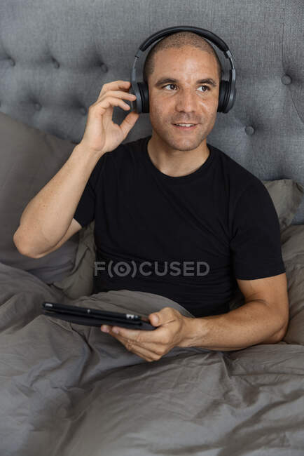 Hombre sereno en auriculares sentados en la cama debajo de la manta y navegando por las redes sociales en la tableta mientras escucha música por la mañana - foto de stock